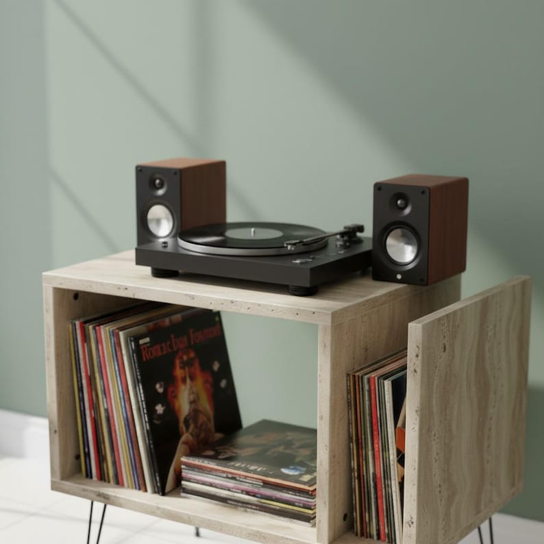 May include: A black turntable with a record on top, flanked by two brown and black speakers. The setup sits on a light-colored wooden cube shelf, with vinyl records visible inside. The background is a solid, muted green.