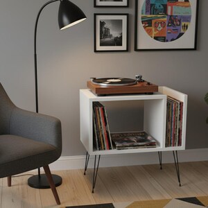 May include: A white record player stand with black hairpin legs. A wooden record player sits on top. The stand holds vinyl records. A black floor lamp and a gray armchair are in the background. Artwork hangs on the wall.