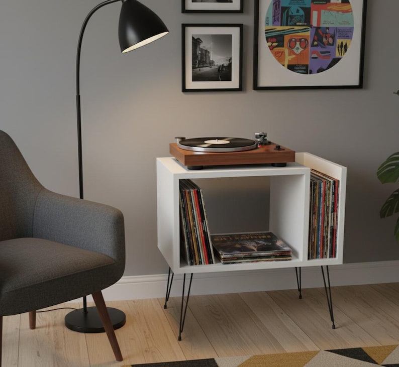 May include: A white record player stand with black hairpin legs. A wooden record player sits on top. The stand holds vinyl records. A black floor lamp and a gray armchair are in the background. Artwork hangs on the wall.