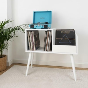 Puede incluir: Un mueble blanco para guardar discos de vinilo con un tocadiscos turquesa. La unidad tiene tres secciones para discos y se apoya en patas blancas en ángulo. Una planta en maceta está en el fondo.