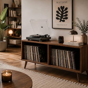 May include: A dark wood media console with two open storage compartments filled with vinyl records. A black turntable with a clear dust cover sits on top, alongside a small candle, a metal lamp, and two stacked books.