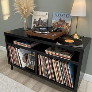 Könnte beinhalten: Eine schwarze Schallplattenkonsole mit einem hölzernen Plattenspieler, einer Lampe und mehreren Schallplatten. Die Konsole hat zwei Regale, die mit Alben gefüllt sind. Eine Vase mit getrockneten Blumen steht oben drauf. Der Raum hat ein neutrales Farbschema.