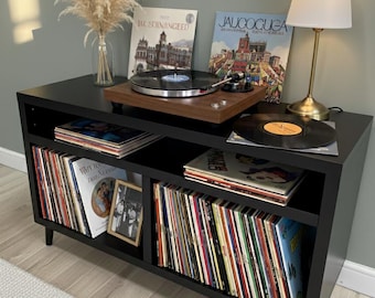 May include: A black record console with a wooden turntable, a lamp, and several vinyl records. The console has two shelves filled with albums. A vase of dried flowers sits on top. The room has a neutral color scheme.