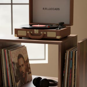 May include: A wooden record player with a black vinyl record playing. The record player is brown with gold accents and a handle. The text "R.R.LUCCADS" is printed on the inside of the lid. A stack of vinyl records and black headphones are in the foreground.