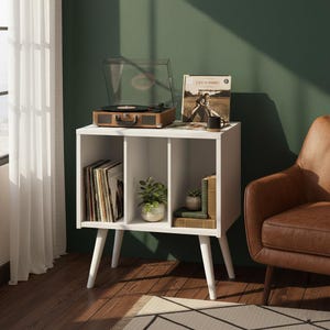 May include: A white mid-century modern record player stand with a brown record player on top. The stand has three cubby holes, filled with vinyl records, plants, and books. The stand is on white angled legs.