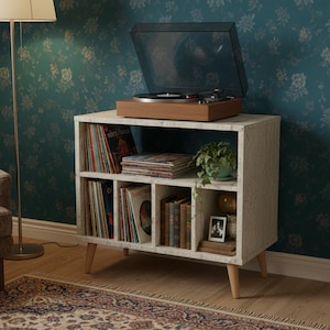 Può includere: Un giradischi vintage su un mobile bianco con gambe in legno. Il mobile è pieno di dischi in vinile, libri e una piccola pianta in vaso. Il giradischi è aperto, pronto all'uso.