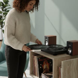May include: A person holding a vinyl record near a black turntable with two brown speakers. The turntable is on a light-colored wooden cabinet filled with vinyl records. The person is wearing a cream-colored sweater and black jeans.