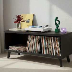 May include: A black record player stand with a white turntable, vinyl records, and decorative items. The stand has angled legs and open shelving for record storage. A small red plant in a silver pot sits on top.