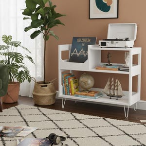 May include: A white bookshelf in a mid-century modern style, with a record player, books, and decorative items. The unit has three shelves and hairpin legs. A Led Zeppelin IV album is visible. A globe and model sailboat are also on display.