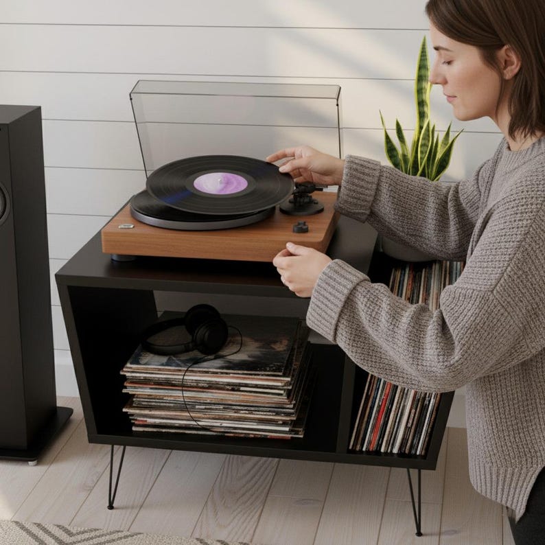 May include: A wooden record player with a clear cover sits on a black cabinet with hairpin legs. A person is placing a vinyl record on the turntable. A stack of vinyl records and a pair of black headphones are visible.