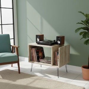 May include: A light wood-grain record player stand with black hairpin legs. The stand holds a black turntable, two small speakers, and vinyl records. A teal chair and a potted plant are in the background.