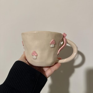 Könnte beinhalten: Eine beige Keramiktasse mit einem skurrilen Design. Die Tasse hat drei rosa Pilzapplikationen und ein rosa Schleifendetail am Griff. Die Tasse wird in einer Hand gehalten, um ihre Größe und Form zu zeigen.