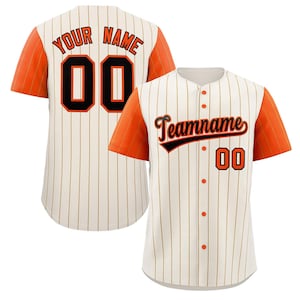 Könnte beinhalten: Cremefarbenes Baseball-Trikot mit orangefarbenen Ärmeln und Nadelstreifen. Das Trikot zeigt den Text "YOUR NAME" und die Nummer "00" auf dem Rücken sowie "Teamname" und "00" auf der Vorderseite. Das Trikot hat orangefarbene Knöpfe.
