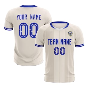 May include: Two cream-colored soccer jerseys with blue accents. One jersey displays "YOUR NAME" and "00" on the back. The other jersey shows "TEAM NAME" and "00" on the front. Both jerseys have a blue collar and sleeve trim.