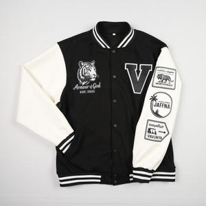 May include: Black and white varsity jacket with white sleeves. The jacket features a tiger graphic and the text "Armour of God EST. 2025" on the chest. Patches on the right sleeve include a tiger, palm tree, and text.