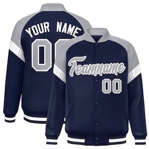 May include: Navy blue and gray varsity jacket with white accents. The jacket features the text "YOUR NAME" and the number "0" on the back, and "Teamname" and the number "00" on the front. The jacket has a button-up front and striped cuffs.