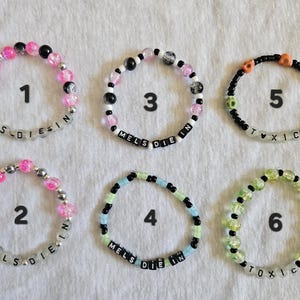 May include: Six beaded bracelets in various colors are displayed. The bracelets feature a mix of clear, black, pink, green, and blue beads. Some bracelets spell out "MELS DIE IN" and "TOXIC" with black letter beads. The bracelets are on a white surface.