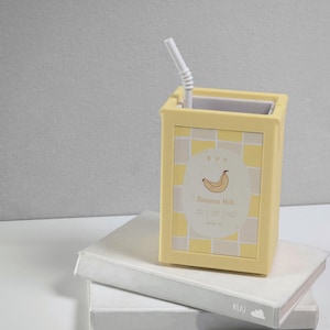Op de afbeelding: Een gele container met een wit rietje, ontworpen om op een pak bananenmelk te lijken. De container heeft een dambordpatroon en een label met de tekst "Banana Milk" met een bananenillustratie. De container staat op twee witte boeken.