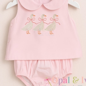 May include: Pink baby outfit set with a collared top and bloomers. The top features embroidered geese with pink bows. The outfit is displayed on a white hanger.