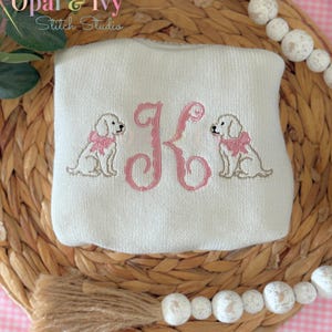 May include: A white, square-shaped baby blanket with an embroidered pink letter "K" in the center. Two embroidered puppies wearing pink bows flank the letter. The blanket is from Opal & Ivy Stitch Studio.