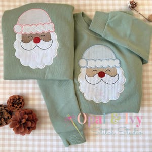 May include: Two sage green sweatshirts with a Santa Claus applique. The Santa design features a white beard, a red nose, and a white hat with pink trim. The sweatshirts are displayed on a checkered surface, with pinecones in the background.