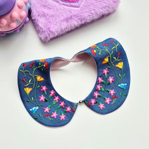 May include: A denim collar with embroidered flowers in shades of pink, yellow, and blue. The collar has a light pink lining and a silver clasp. A purple fuzzy item and a pink toy are in the background.
