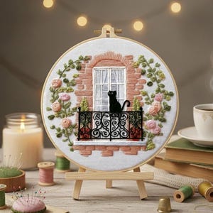 以下が含まれることがあります： 黒猫がバルコニーにいる刺繍フープアート。レンガ造りの窓が特徴です。デザインにはピンクのバラと緑の葉が含まれています。このアートワークは小さな木製イーゼルに展示されています。