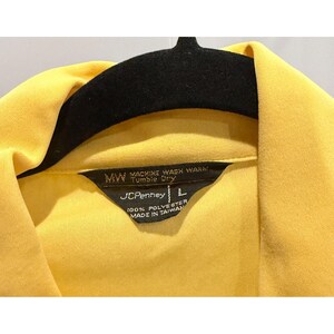 May include: Close-up of a yellow polyester shirt with a black label. The label reads "MACHINE WASH WARM, TUMBLE DRY" and "JCPenney L". The shirt is hanging on a black hanger.