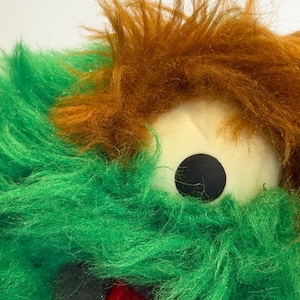 May include: Close-up of a plush toy with green and brown shaggy fur. The toy has a large, round, white eye with a black pupil. A red, fuzzy mouth is visible at the bottom of the image. The toy appears to be a monster or creature.