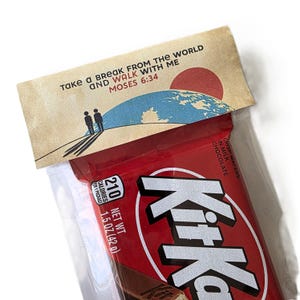 Puede incluir: Una barra de chocolate Kit Kat roja en un envoltorio transparente, con una tarjeta decorativa encima. La tarjeta presenta un mapa del mundo, un sol rojo y el texto "Take a break from the world and walk with me Moses 6:34". La barra pesa 42 g.