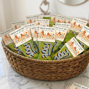 Pu&ograve; includere: Un cesto intrecciato riempito con diversi sacchetti di snack verdi al gusto di frutta. Ogni sacchetto presenta un'intestazione in cartoncino bianco stampata con il testo "WALK WITH ME" "ONE FOOT AT A TIME" "MOSES 6:34" e un'illustrazione di piedi e fiori.