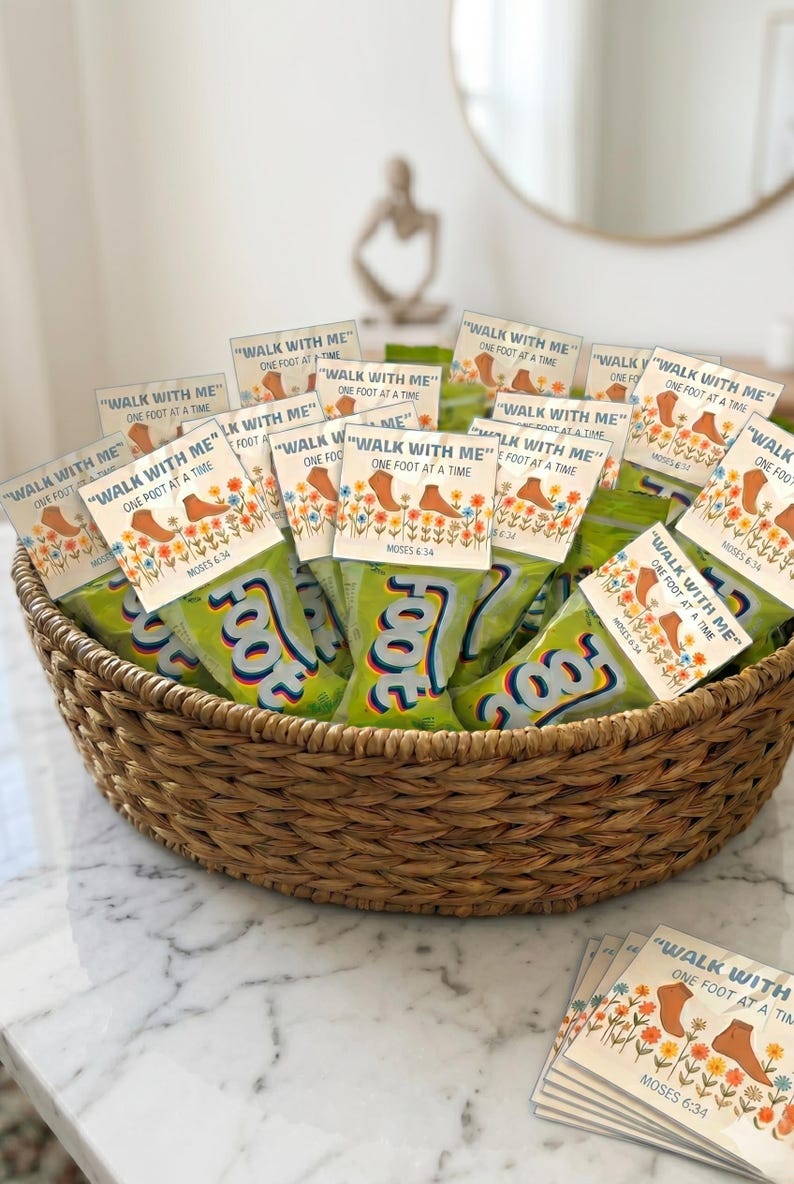 Pu&ograve; includere: Un cesto intrecciato riempito con diversi sacchetti di snack verdi al gusto di frutta. Ogni sacchetto presenta un'intestazione in cartoncino bianco stampata con il testo "WALK WITH ME" "ONE FOOT AT A TIME" "MOSES 6:34" e un'illustrazione di piedi e fiori.