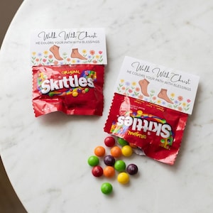 Peut inclure: Deux sachets de bonbons Skittles rouges avec des étiquettes en papier blanc portant le texte Walk With Christ HE COLORS YOUR PATH WITH BLESSINGS, des illustrations de pieds et des fleurs colorées. Un sachet est ouvert avec des bonbons.