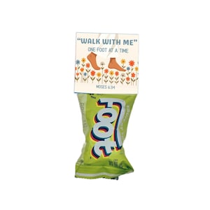 Pu&ograve; includere: Una confezione di snack verde chiaro etichettata "Fruit by the Foot" con un topper di carta rettangolare che riporta il testo "WALK WITH ME" e "ONE FOOT AT A TIME" sopra un'illustrazione di due piedi circondati da fiori colorati e "MOSES 6:34".