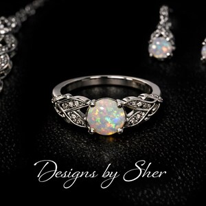 Könnte beinhalten: Ein Schmuckset mit Ring, Ohrringen und Halskette, alle in Silber. Ring und Ohrringe haben einen zentralen Opalstein, der Ring ein dekoratives Blattdesign. Der Text "Designs by Sher" ist unten zu sehen.