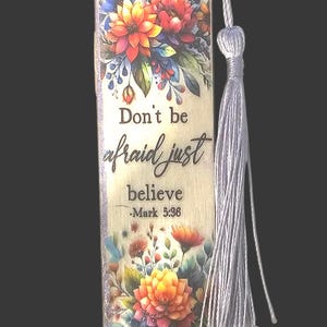 May include: A rectangular wooden bookmark featuring a vibrant floral print with orange, red, and blue flowers. The text reads, "Don't be afraid just believe -Mark 5:36." A white cord tassel is attached to the top hole of the bookmark.