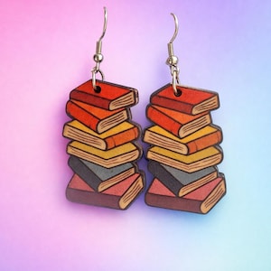 Puede incluir: Pendientes con forma de pilas de libros coloridos, con tonos de rojo, naranja, amarillo y gris. Los pendientes tienen ganchos plateados. El texto "Designs by Sher" es visible en la parte superior. Un accesorio encantador para los amantes de los libros.