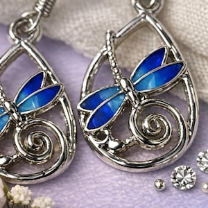 Può includere: Orecchini a forma di libellula color argento con ali in smalto blu. Le montature a forma di goccia presentano un design a libellula con dettagli intricati e un accento a spirale. Cristalli e perle scintillanti sono sparsi intorno agli orecchini.