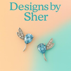 May include: A pair of silver hummingbird earrings, each with a light blue, teardrop-shaped gemstone. The wings are embellished with small, clear crystals. The image includes the text "Designs by Sher" in teal.