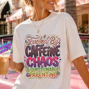 May include: A white short-sleeve t-shirt featuring a colourful graphic print with the text "Fueled by CAFFEINE CHAOS & QUESTIONABLE PARENTING" in various fonts and styles, including small illustrations of coffee cups and a calendar icon.