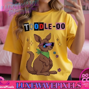 Può includere: T-shirt gialla con la scritta "TOODLE-OO" in un design a blocchi colorati. Sul davanti è raffigurato un canguro dei cartoni animati con occhiali da sole e una sciarpa verde, insieme a stelle e fiori.