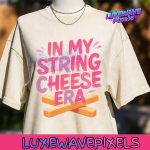 Op de afbeelding: Crèmekleurig T-shirt met de tekst "IN MY STRING CHEESE ERA" in roze en paars. Twee oranje kaasstengels zijn onder de tekst gekruist. Een gouden ketting is zichtbaar.