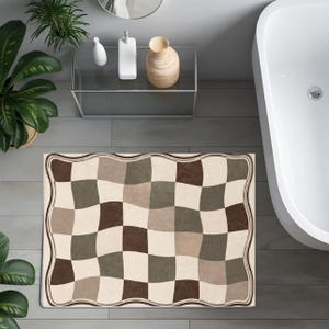 Puede incluir: Una alfombrilla de baño rectangular con un patrón de tablero de ajedrez ondulado y distorsionado en tonos crema, beige y marrón oscuro. La alfombrilla tiene un borde decorativo ondulado que sigue los contornos curvos del diseño.