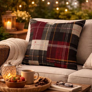 May include: A square cushion with a patchwork plaid pattern in burgundy, black, grey, and beige. The cushion is on a beige sofa, creating a cosy atmosphere. The cushion is a home decor item. The cushion is approximately 40cm x 40cm.