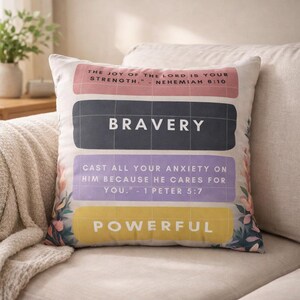 Könnte beinhalten: Quadratisches Zierkissen mit inspirierenden Zitaten. Das Kissen zeigt die Wörter "BRAVERY" und "POWERFUL" in großem Text, zusammen mit anderen Sätzen und floralen Akzenten. Der Hintergrund ist neutral gefärbt.