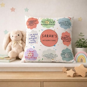 Puede incluir: Un cojín cuadrado blanco con afirmaciones coloridas, incluyendo "Soy digno de amor y felicidad." El cojín tiene un círculo central con "SARAH'S AFFIRMATIONS." Un conejito de peluche y estrellas de madera están cerca.