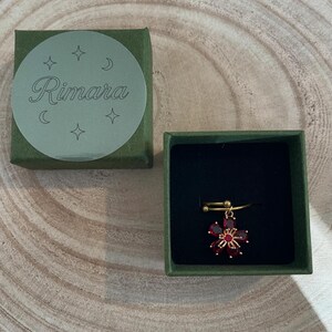 Könnte beinhalten: Ein goldfarbener Ring mit einem hängenden roten Blumenanhänger mit facettierten Blütenblättern, präsentiert in einer quadratischen dunkelgrünen Schachtel. Der Deckel zeigt einen runden Aufkleber mit dem Text Rimara und Stern- sowie Mondsymbolen.