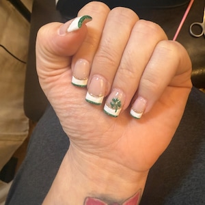 May include: A hand with manicured nails featuring a French tip design with white, green glitter, and gold accents. One nail has a green shamrock design. The nails are short and square-shaped.