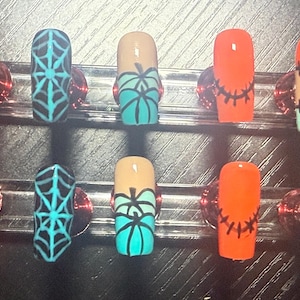 May include: A set of press-on nails with a Halloween theme. Designs include orange nails with black stitches, teal spiderwebs, pumpkin designs, and tan nails with patchwork. The nails are displayed on a clear stand.