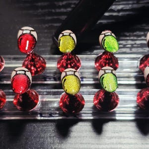 Peut inclure: Un ensemble de faux ongles avec un motif d'œil de dessin animé. Les ongles présentent une base blanche avec des motifs d'yeux rouges, jaunes et verts. Chaque ongle est monté sur un présentoir rectangulaire transparent avec des accents sphériques rouges.